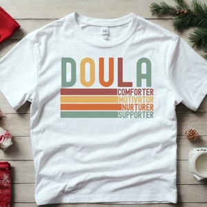 Op de afbeelding: Wit T-shirt met het woord "Doula" in een retro-lettertype met kleurrijke strepen eronder. De strepen hebben de woorden "trooster", "motivator", "verzorger" en "ondersteuner" erop.