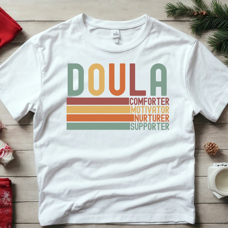 Op de afbeelding: Wit T-shirt met het woord "Doula" in een retro-lettertype met kleurrijke strepen eronder. De strepen hebben de woorden "trooster", "motivator", "verzorger" en "ondersteuner" erop.