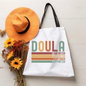 Op de afbeelding: Witte canvas tote bag met zwarte handvatten. De tas heeft het woord "DOULA" in kleurrijke retro-stijl letters gedrukt. Onder het woord staat de tekst "COMFORTER, MOTIVATOR, NURTURER, SUPPORTER" in kleinere letters.