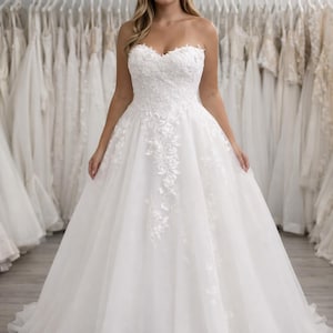 Op de afbeelding: Een strapless witte trouwjurk met een sweetheart halslijn en bloemen kanten details. De volle rok is gemaakt van lagen tule. De jurk wordt getoond in een bruidszaak met andere jurken op de achtergrond.