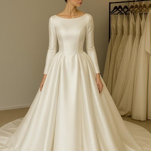 Op de afbeelding: Een ivoorkleurige trouwjurk met een boothals en lange mouwen. De jurk heeft een getailleerd lijfje en een volle rok die uitloopt in een sleep. De satijnen stof heeft een subtiele glans. Andere jurken hangen op de achtergrond.