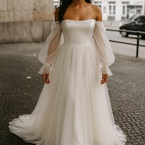 Op de afbeelding: Een witte off-the-shoulder trouwjurk met lange, doorschijnende mouwen en een zwierige tule rok. De jurk heeft een getailleerd lijfje en een sleep. De bruid staat op een geplaveide straat.