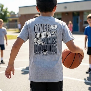 Puede incluir: Una camiseta gris con el texto "OVERTHINKING SOLVES NOTHING" e ilustraciones de robots. La persona lleva un balón de baloncesto. El texto también incluye "BUT ACTION BRINGS CLARITY".