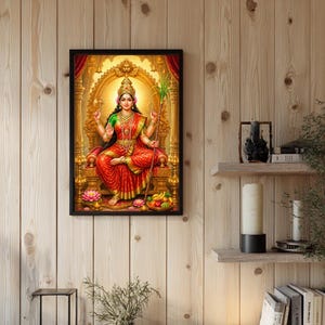May include: A framed wall art print depicting a deity seated on an ornate golden throne. The figure wears a red and green sari with gold jewelry, holding a sugarcane stalk and a lotus. A bowl of fruit and pink lotus flowers sit at the base.