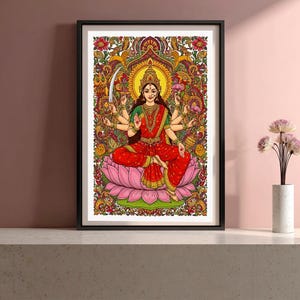 Puede incluir: Ilustración enmarcada y colorida de una deidad de múltiples brazos sentada sobre una flor de loto rosa. La deidad lleva un sari rojo y una blusa verde, con un halo y una corona dorados. El fondo está lleno de intrincados patrones florales.