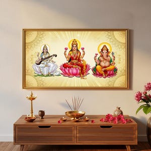 Könnte beinhalten: Ein gerahmtes Kunstwerk mit drei hinduistischen Gottheiten: Saraswati, Lakshmi und Ganesha. Die Figuren sind in leuchtenden Farben dargestellt und sitzen auf Lotusblumen vor einem goldenen Hintergrund. Das Kunstwerk wird über einem Holzschrank ausgestellt.