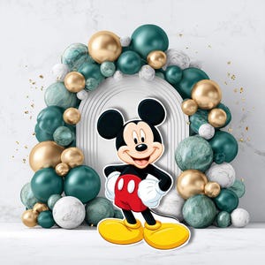 May include: A cheerful Mickey Mouse cutout stands before an arch of teal, gold, and marble-textured balloons. The iconic character is dressed in red shorts, yellow shoes, and white gloves, smiling brightly. The background features a white arch and gold confetti.