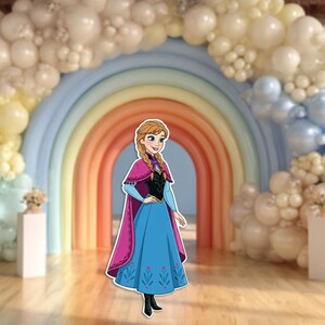 May include: A cutout of a cartoon character in a blue dress with a pink cape, standing in front of a rainbow arch. The background includes a balloon arch in shades of white, yellow, and blue, creating a festive atmosphere.