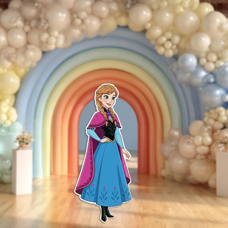 May include: A cutout of a cartoon character in a blue dress with a pink cape, standing in front of a rainbow arch. The background includes a balloon arch in shades of white, yellow, and blue, creating a festive atmosphere.