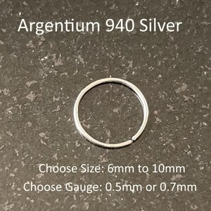 May include: A thin, circular Argentium 940 silver hoop ring. The text reads: Argentium 940 Silver, Choose Size: 6mm to 10mm, Choose Gauge: 0.5mm or 0.7mm. The silver ring is a simple, continuous band with a small gap, showcasing a metallic finish.