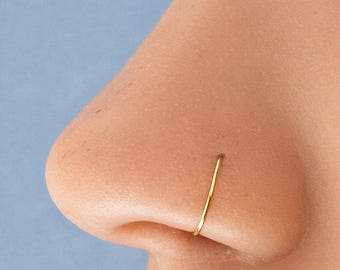 May include: A thin, circular gold nose ring hoop worn in the nostril. The jewelry features a smooth, polished metallic finish and a simple, continuous design that rests flush against the skin of the nose, creating a subtle and refined appearance.