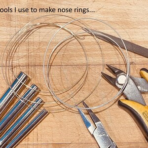 May include: A collection of jewelry-making tools including four silver-toned metal mandrels, a pair of yellow and black handled wire cutters, a pair of needle-nose pliers, a metal file, and coiled silver wire. Text reads: The tools I use to make nose rings...
