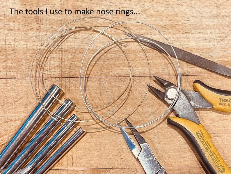 May include: A collection of jewelry-making tools including four silver-toned metal mandrels, a pair of yellow and black handled wire cutters, a pair of needle-nose pliers, a metal file, and coiled silver wire. Text reads: The tools I use to make nose rings...
