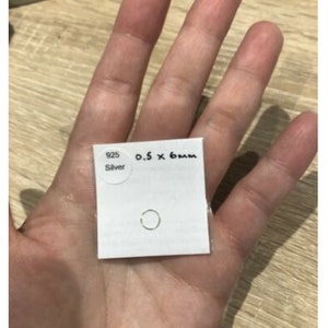 May include: A small silver ring on a white square card. The card features a circular sticker with the text 925 Silver and handwritten text reading 0.5 x 6mm. The ring is a thin, circular band made of 925 sterling silver, measuring 0.5mm by 6mm.