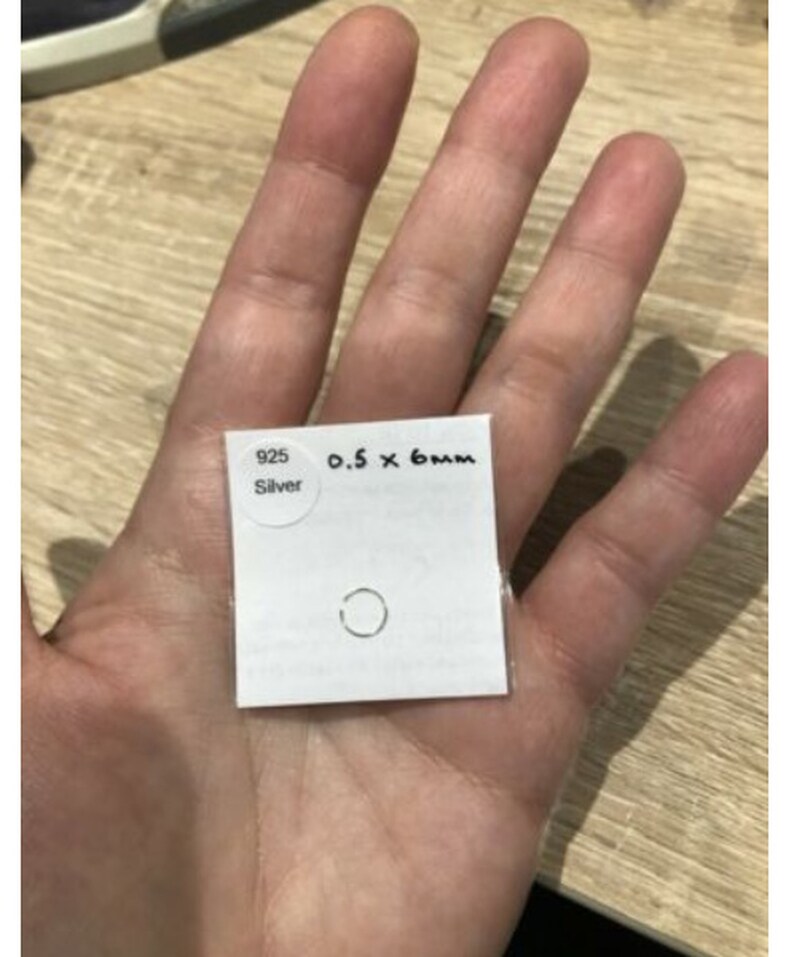 May include: A small silver ring on a white square card. The card features a circular sticker with the text 925 Silver and handwritten text reading 0.5 x 6mm. The ring is a thin, circular band made of 925 sterling silver, measuring 0.5mm by 6mm.