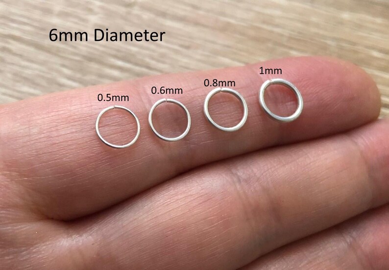 May include: Four silver-colored metal jump rings with a 6mm diameter, displayed in varying wire thicknesses labeled 0.5mm, 0.6mm, 0.8mm, and 1mm. These circular findings are arranged in a row, showcasing the different gauge options available for jewelry making.