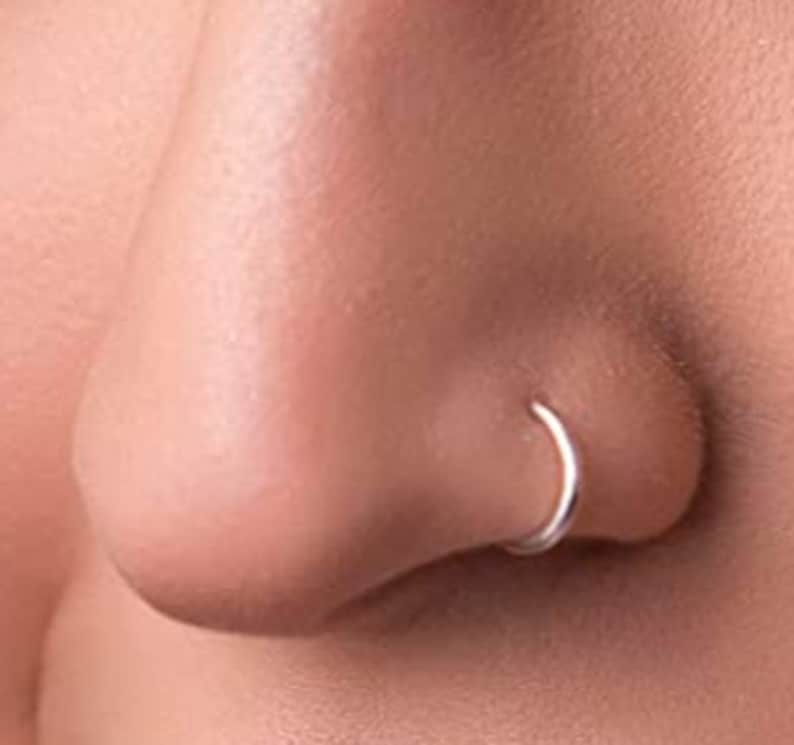 May include: A thin, circular silver nose ring hoop positioned in the nostril. The jewelry features a smooth, polished metallic finish and a simple, continuous band design that forms a complete circle when worn in the nasal piercing.
