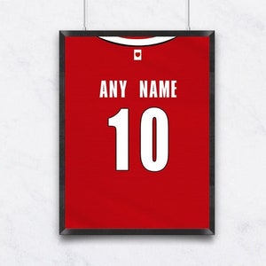 Puede incluir: Camiseta roja enmarcada con el texto "ANY NAME" encima del número "10" en blanco. La camiseta se exhibe en una pared blanca, suspendida por dos finos cordones blancos.