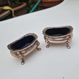 以下が含まれることがあります： ミニチュアの浴槽の形をした、2つのアンティークシルバーのソルトセラー。各浴槽の内部は深く光沢のある黒色です。ソルトセラーは装飾的なディテールがあり、小さな湾曲した足の上に置かれ、ヴィンテージの魅力を添えています。