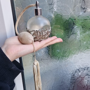 Puede incluir: Un frasco de perfume vintage con una base de metal plateado y un cuerpo de vidrio esmerilado. El frasco tiene un atomizador de bulbo y una borla larga. La base está decorada con diseños florales. El frasco se sostiene en una mano.