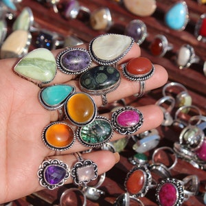 Puede incluir: Una colección de anillos de piedras preciosas con varias formas y colores, incluyendo turquesa, ámbar y amatista. Los anillos tienen bandas de metal plateado y se exhiben sobre una superficie de madera.