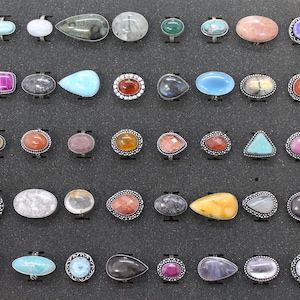 May include: A collection of gemstone rings displayed on a black surface. The rings feature various shapes and colors, including red, blue, green, and purple stones. Each ring has a silver-colored band.
