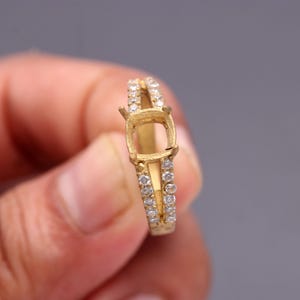 Puede incluir: Un engaste de anillo de color dorado con un diseño de vástago dividido. El anillo presenta un engaste cuadrado para una piedra preciosa y está adornado con pequeñas piedras transparentes a lo largo de la banda. El anillo se sostiene sobre un fondo gris.
