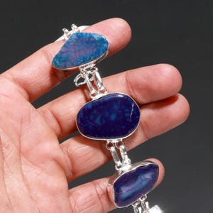 Puede incluir: Una pulsera de plata con tres piedras grandes de forma irregular. Las piedras son de color azul oscuro y morado con patrones intrincados. La pulsera se sostiene en una mano sobre un fondo oscuro.