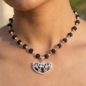 May include: A necklace with dark brown beads and a silver pendant. The pendant displays the word "महाकाल" within a heart-shaped frame. The beads are connected with silver links, forming a detailed and elegant piece of jewellery.