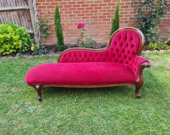 May include: A red velvet chaise lounge with a tufted back and wooden legs. The chaise lounge is in a garden setting.