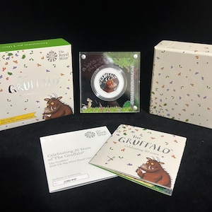 May include: A collection of items celebrating 20 years of "The Gruffalo." This includes a 2019 UK 50p Silver Proof Coin, displayed in a clear case, with packaging and a certificate of authenticity. The coin features the Gruffalo character.
