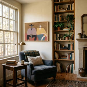May include: A framed rectangular wall art piece depicting two stylized figures in a hat and yellow top, displayed above a dark gray armchair with a plaid throw, a wooden side table with books, and a tall bookshelf filled with various books.
