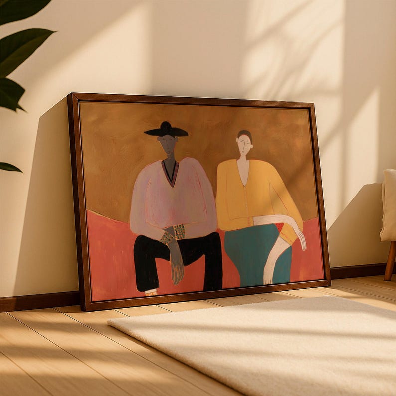 May include: A framed rectangular wall art print featuring a stylized illustration of two figures sitting side by side. The figure on the left wears a black hat, a light pink long-sleeved top, and black pants. The figure on the right wears a yellow long-sleeved top and a teal skirt, set against a warm brown and red background.