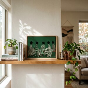 May include: A framed art print featuring five figures in black and white striped clothing against a green background. The artwork is displayed on a wooden shelf with books, plants, and a candle holder. The scene is set in a well-lit interior.