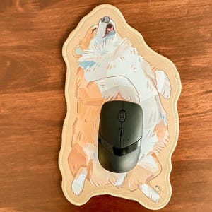 May include: A die-cut mouse mat featuring a tan and white illustration of a dog lying on its back with its paws in the air. A black wireless computer mouse with a scroll wheel and two side buttons is positioned in the centre of the mouse mat.