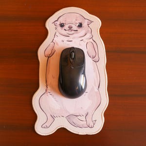 Puede incluir: Un ratón de ordenador negro descansa sobre una alfombrilla con una ilustración de un chihuahua marrón claro. La alfombrilla tiene un borde crema y está sobre una superficie de madera marrón oscuro.