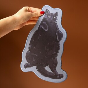 May include: A black cat illustration on a light blue, irregular-shaped patch. The cat is depicted in a playful pose, with its paws raised. The patch is held against a brown background, with a green plant and a person's arm and wrist with a bracelet visible.