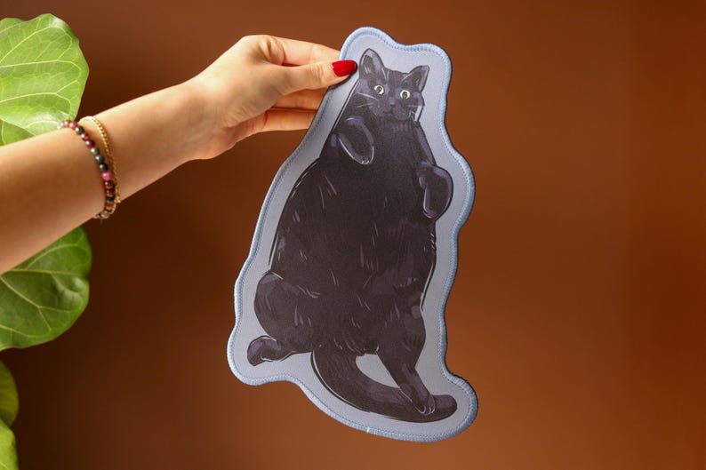 May include: A black cat illustration on a light blue, irregular-shaped patch. The cat is depicted in a playful pose, with its paws raised. The patch is held against a brown background, with a green plant and a person's arm and wrist with a bracelet visible.
