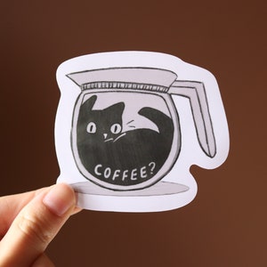 Op de afbeelding: Een witte sticker met een zwart-witte illustratie van een koffiekan met een zwarte kat erin. De tekst "COFFEE?" staat onderaan de koffiekan.