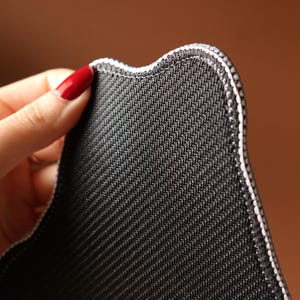 May include: Close-up of a black, textured fabric with a white stitched border. The fabric has a subtle diagonal weave pattern. The edge of the fabric has a curved shape. The background is a blurred brown.