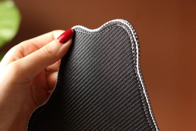 May include: Close-up of a black, textured fabric with a white stitched border. The fabric has a subtle diagonal weave pattern. The edge of the fabric has a curved shape. The background is a blurred brown.