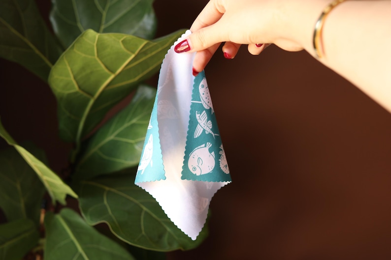May include: A teal blue fabric kitchen towel with a white border and a white print of various sea creatures. The towel is folded in half and held by a hand.