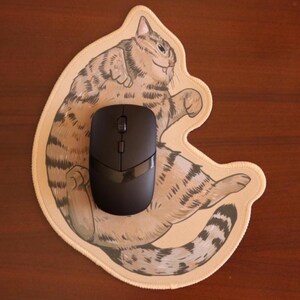 May include: A black wireless computer mouse sits on a beige mouse mat shaped like a cartoon cat. The cat illustration has brown and grey stripes and a smiling face. The mouse has a sleek, modern design.