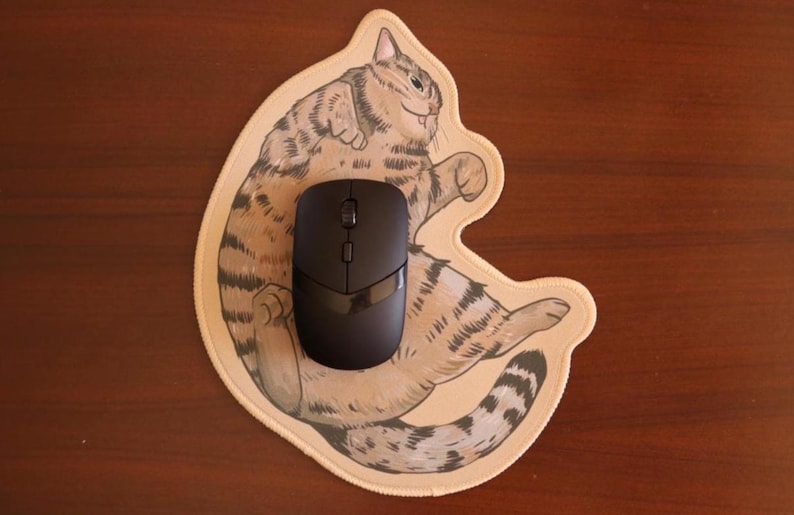 May include: A black wireless computer mouse sits on a beige mouse mat shaped like a cartoon cat. The cat illustration has brown and grey stripes and a smiling face. The mouse has a sleek, modern design.
