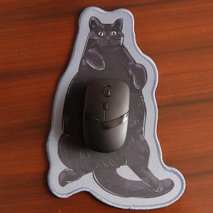 May include: A black computer mouse sits on a mouse mat featuring a black cat design. The mouse mat has a light blue border and a grey cat illustration. The background is a wooden surface.