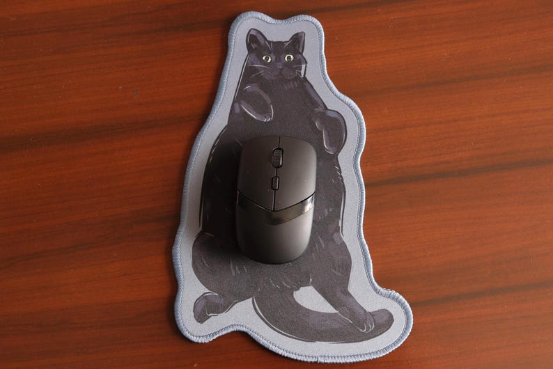 May include: A black computer mouse sits on a mouse mat featuring a black cat design. The mouse mat has a light blue border and a grey cat illustration. The background is a wooden surface.
