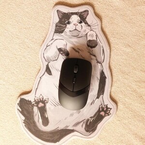 May include: A black wireless computer mouse sits on a cat-shaped mousepad. The mousepad has a cartoon illustration of a black and white cat. The background is a beige, textured surface.