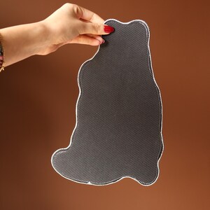 May include: A dark grey, cat-shaped mat with a white stitched border is held up. The mat has a textured surface. The background is a gradient of brown. The hand holding the mat has red nail polish.