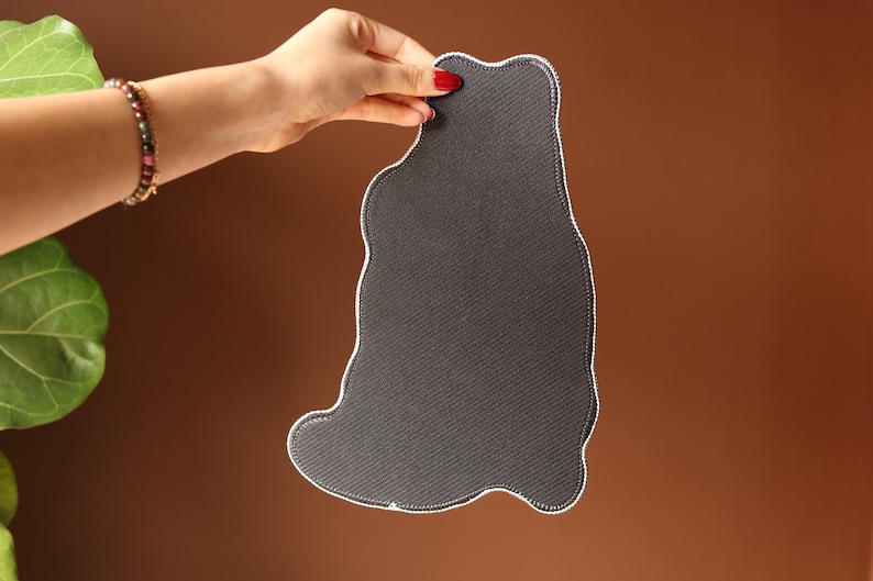 May include: A dark grey, cat-shaped mat with a white stitched border is held up. The mat has a textured surface. The background is a gradient of brown. The hand holding the mat has red nail polish.