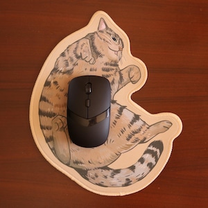 Peut inclure: Un tapis de souris beige avec une illustration de dessin animé d'un chat tigré. Une souris sans fil noire est placée sur le chat.